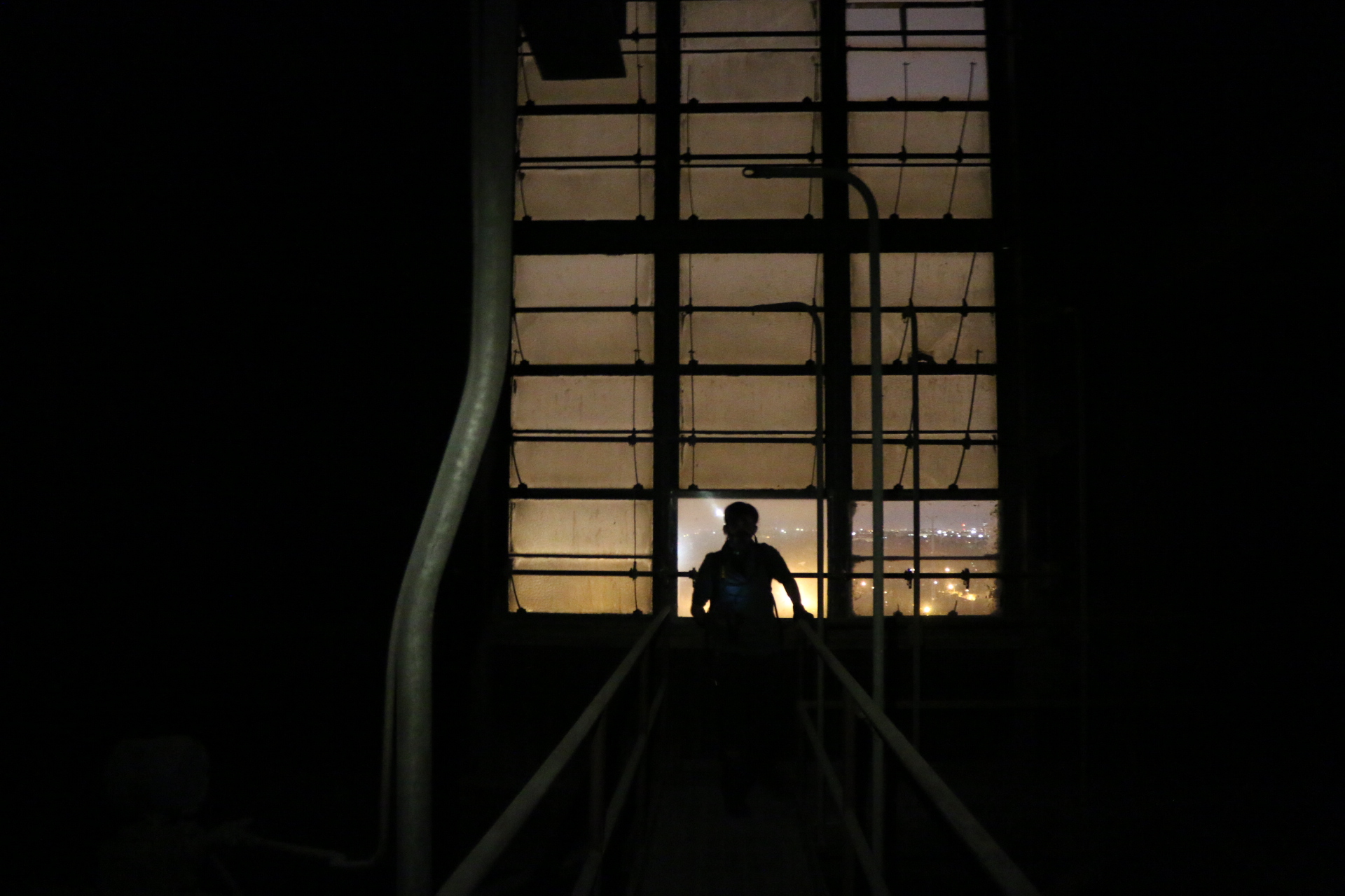 A figure silhouetted at the head of a walkway against a tall grid of industrial windows glowing with sodium light