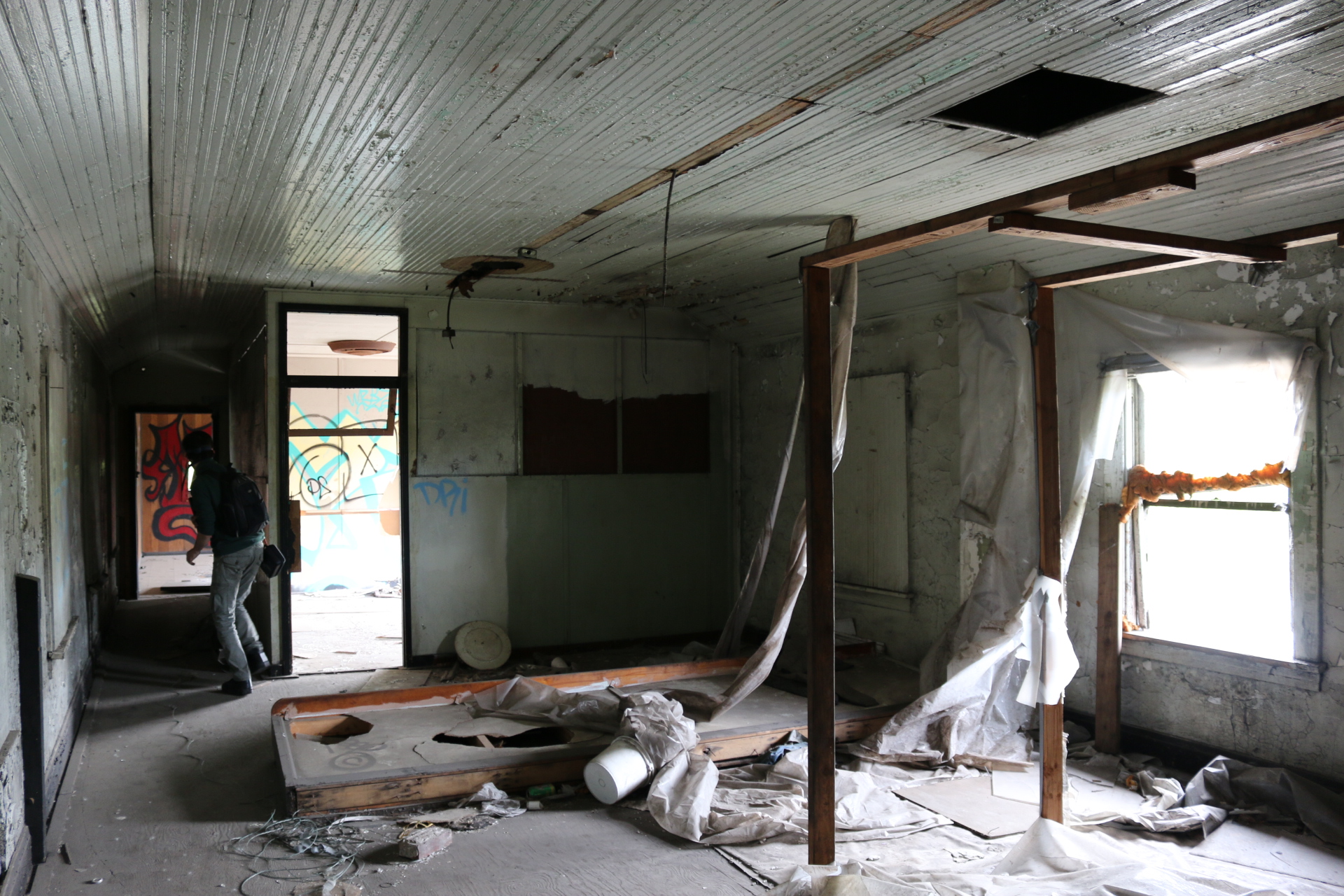 A gutted room with a pressed-tin ceiling and peeling paint, plastic sheeting hanging from a timber frame while a figure cautiously passes through a graffitied doorway
