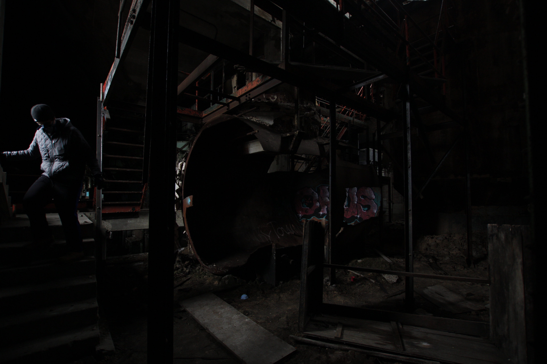 A person climbing a stairway in a dark boiler house beside the riveted end of a large tank