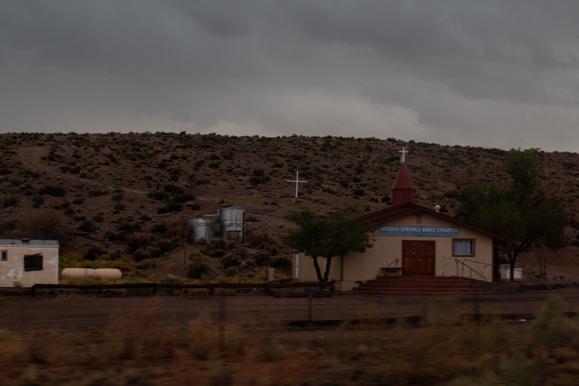 A small roadside church with a red steeple below a scrub-covered hill, a white cross set on the slope behind it
