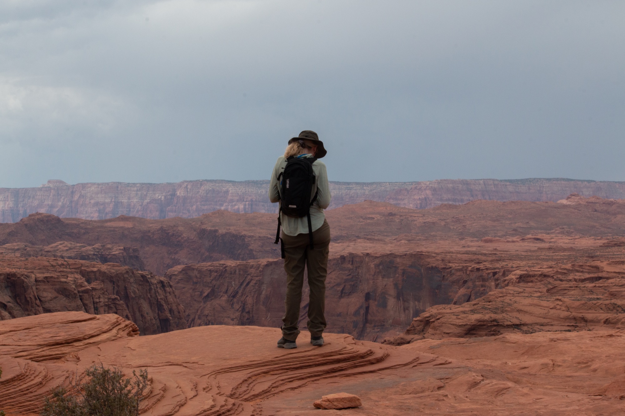 A hiker in a wide-brimmed hat and daypack standing on red slickrock at a canyon rim, looking out toward layered cliffs under a storm sky