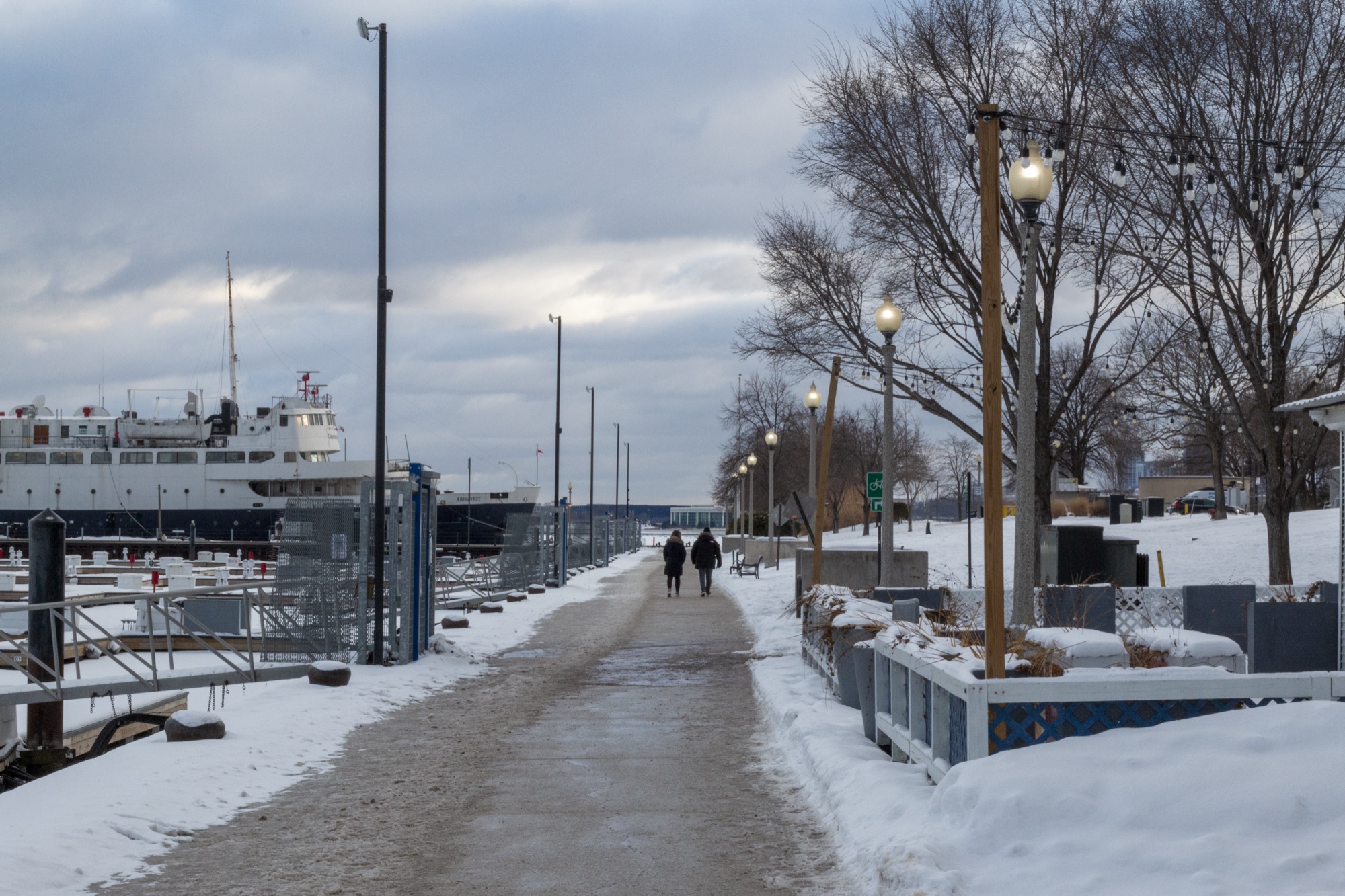 A cleared path along a snowy harbor promenade, lamp posts and bare trees, a large white ship moored to one side, two people strolling together