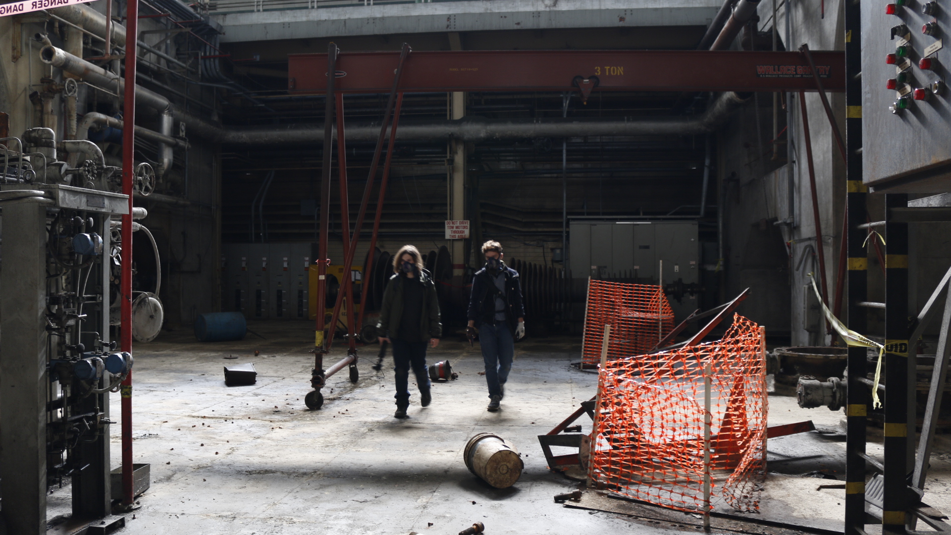 Two people crossing a vacant plant floor past orange safety fencing, an overhead crane rail spanning the ceiling