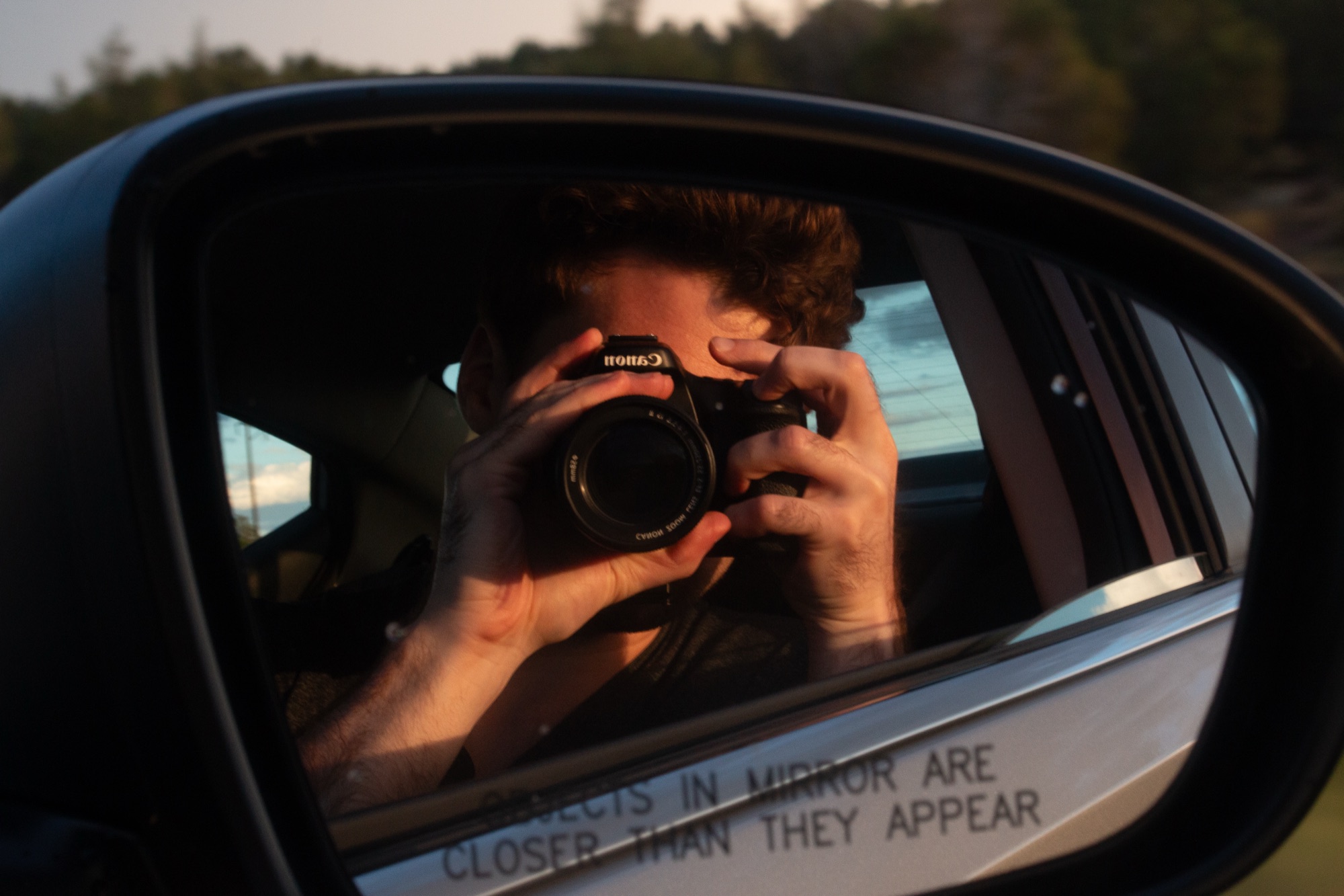 The photographer framed in a car wing mirror raising a Canon DSLR, the words objects in mirror are closer than they appear etched across the glass