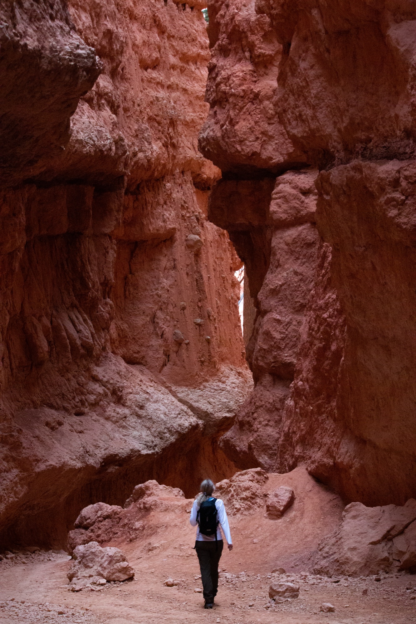 A hiker with a backpack walking a sandy trail into a narrow red rock canyon, the walls rising steeply on either side