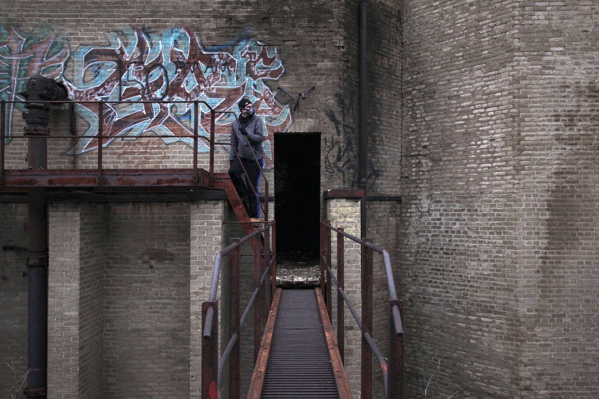 A person on a rusted steel catwalk in a brick shaft, a large blue graffiti piece on the wall behind and a dark doorway ahead