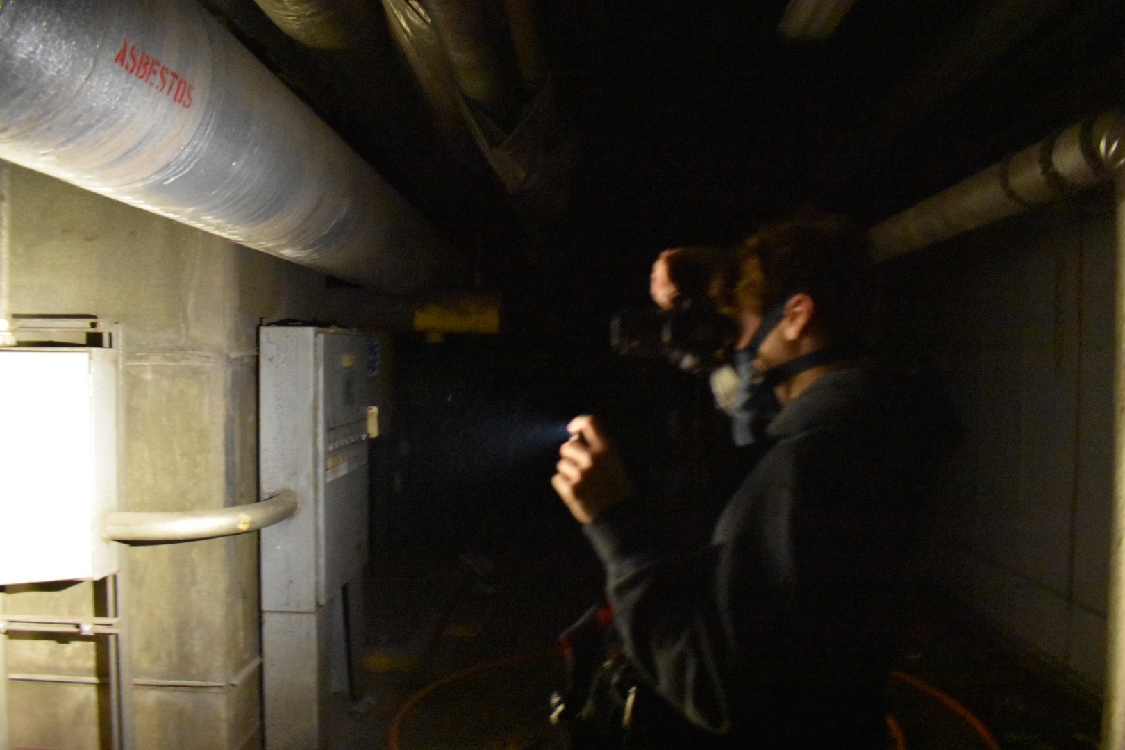 An explorer in a respirator moving down a service tunnel, a torch beam raised toward ducting stenciled ASBESTOS in red