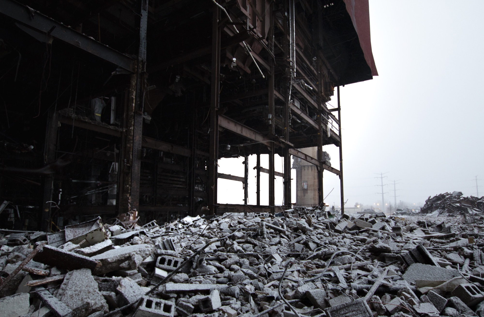 A field of snow-dusted rubble and broken cinder blocks in front of a gutted building stripped back to its steel frame