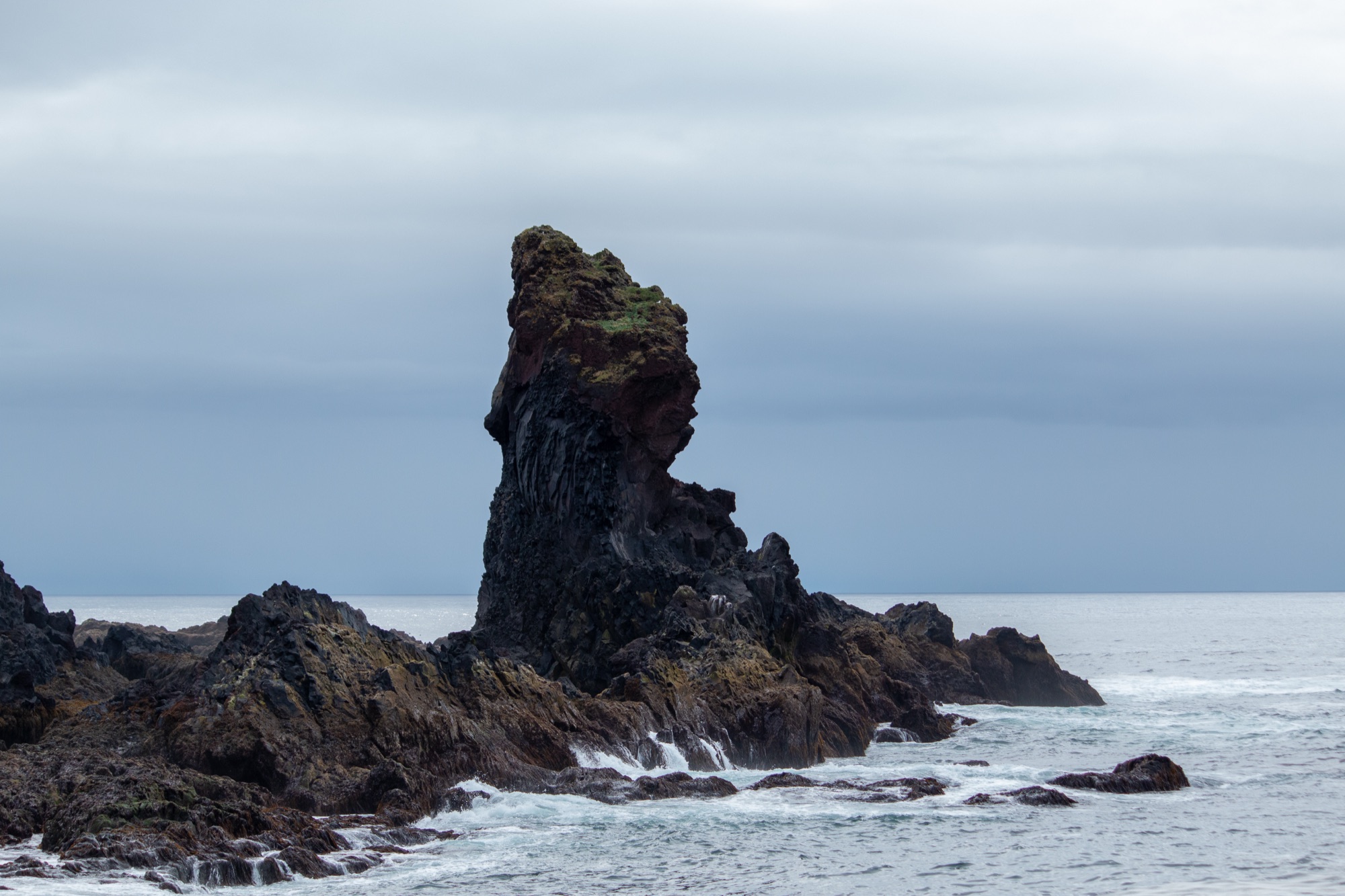 A jagged sea stack of dark lava rock rising from the surf beneath a soft grey sky
