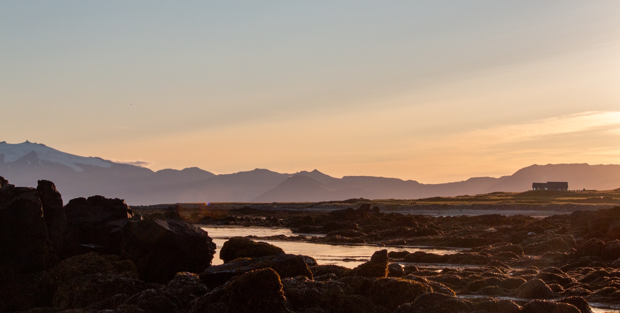 A rocky shoreline at sunset, a glacier-capped mountain range silhouetted across the water