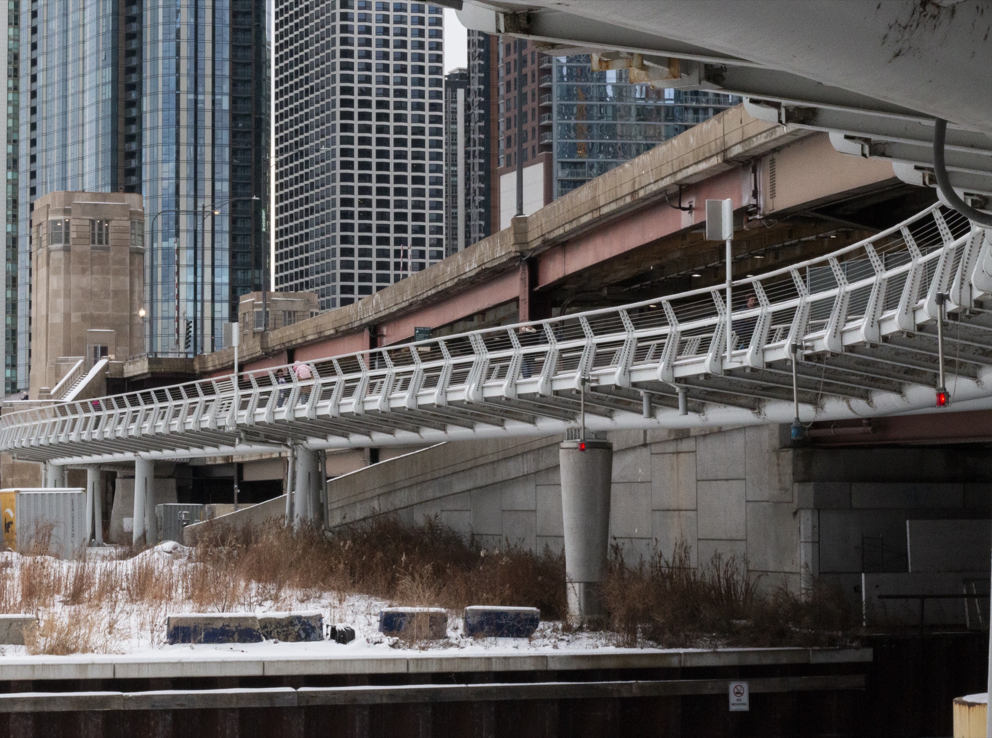 A curving elevated walkway on concrete piers running beneath high-rise towers, snow on the ground below
