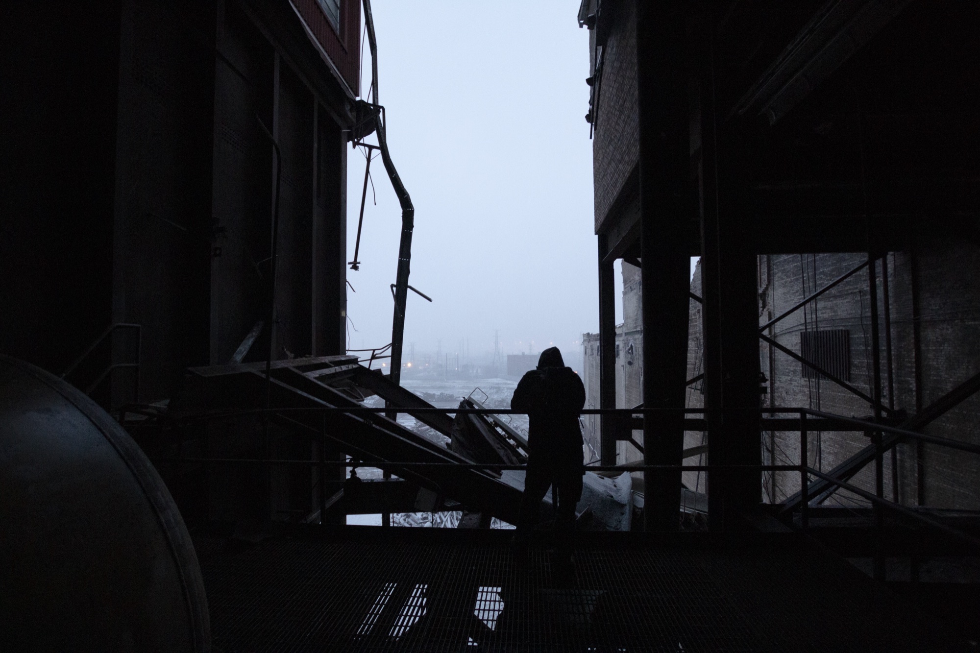 A hooded figure silhouetted on a steel catwalk at the open edge of a derelict industrial building, the skyline beyond lost in a snowy haze